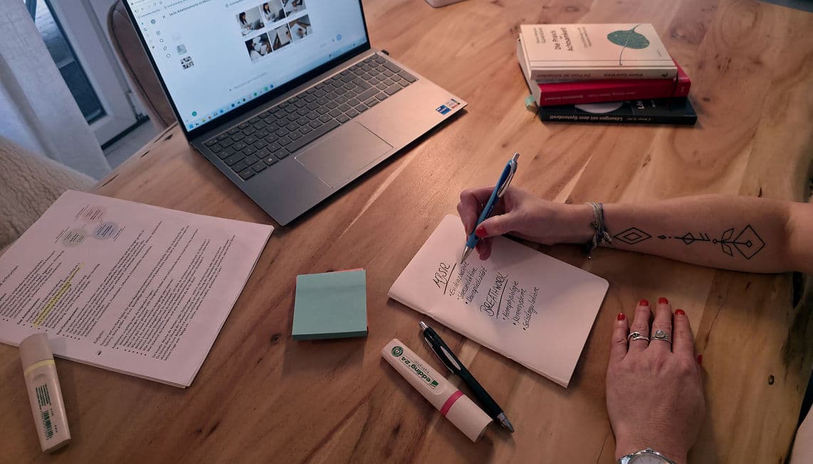 Aufsicht auf einen Holzschreibtisch mit Laptop, markiertem Studienausdruck, mintgrünen Haftnotizen und einer Hand, die Stichworte zu MBSR und Breathwork in ein Notizbuch schreibt