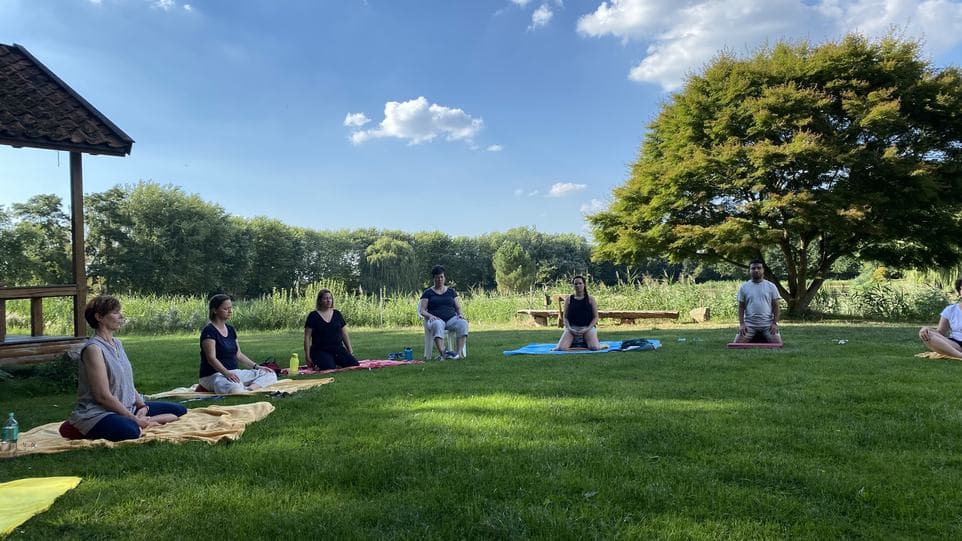 Gruppe sitzt in Meditation auf Decken und Matten auf einer Wiese am Wasser