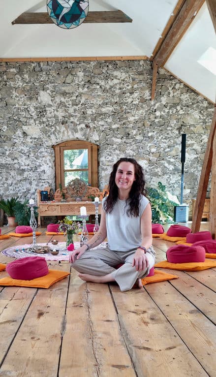 Annika Brüggemann sitzt in einem Raum mit Natursteinwand zwischen vorbereiteten Sitzkissen