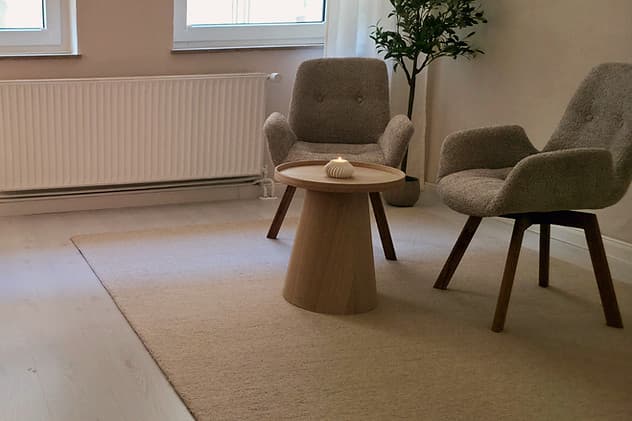 Heller Raum mit Teppich, rundem Holztisch, zwei Sesseln und einem Olivenbaum