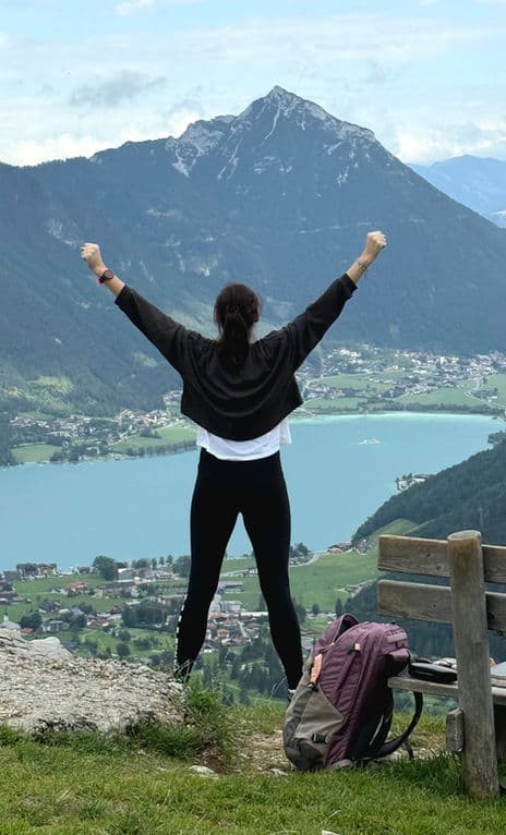 Person mit erhobenen Armen auf einem Berggipfel über einem See